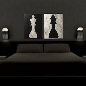 May include: Two black and white silhouette paintings of a king and queen chess piece on a black wall above a dark gray bed with white pillows.