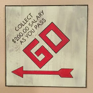 May include: A white painted wooden sign with a red arrow pointing to the right. The sign says "COLLECT $200.00 SALARY AS YOU PASS GO" with the word "GO" in large red letters.