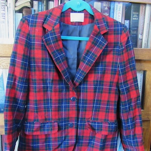 May include: A red, blue, and white plaid blazer with two buttons. The blazer has a pocket on the left side and is hanging on a teal hanger.