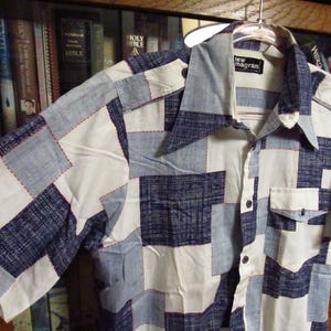 May include: A short-sleeved, white button-down shirt with a blue and white patchwork design. The shirt has a collar and a pocket on the left side. The label on the shirt reads "lew magran".