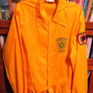 May include: A yellow jacket with a green and white logo that says "Southwest Indian Guide YMCA". The jacket has a patch on the sleeve with a Native American design and the words "Nation YMCA".