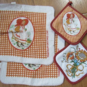 May include: Set of four kitchen towels with a gingham pattern. Two towels feature a girl in a red dress and a red hat, and two towels feature a floral design with a sunflower and a poppy. The towels are white with orange and red gingham patterns. The towels have the text "American Greetings Corp." printed on them.