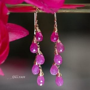 May include: A pair of drop earrings featuring pink, teardrop-shaped gemstones. The gems are wire-wrapped and hang from gold-toned hooks. The earrings are shown near a bright pink flower, creating a vivid contrast.
