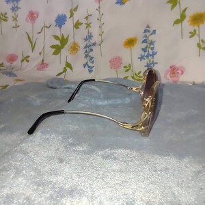 May include: A pair of sunglasses featuring a gold-toned metal frame with a distinct cut-out design at the temples. The arms are thin and metallic, ending in curved black plastic temple tips. The lenses are dark and have a rounded, oversized shape.