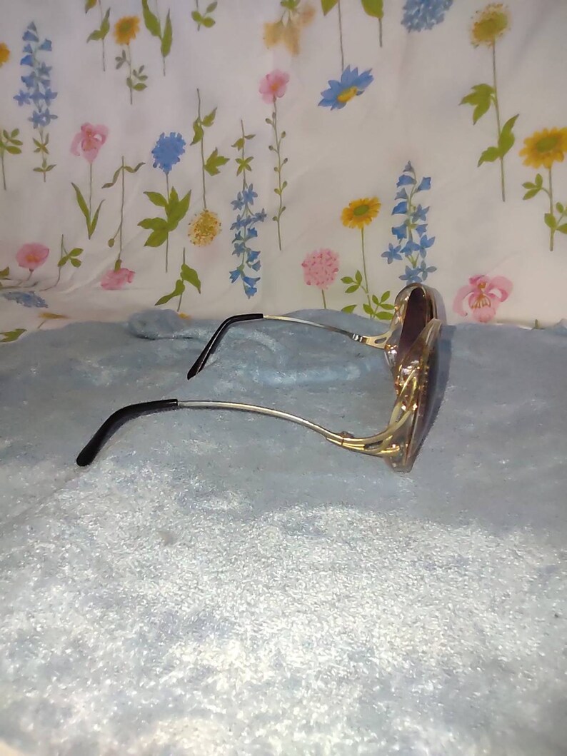 May include: A pair of sunglasses featuring a gold-toned metal frame with a distinct cut-out design at the temples. The arms are thin and metallic, ending in curved black plastic temple tips. The lenses are dark and have a rounded, oversized shape.