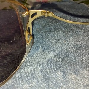 May include: Sunglasses with dark tinted lenses and a gold-toned metal frame. The frame features a decorative cutout design at the hinge where the temple connects to the lens rim. The metal arm is thin and extends outward from the side of the frame.