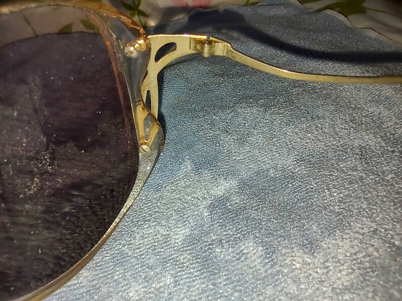 May include: Sunglasses with dark tinted lenses and a gold-toned metal frame. The frame features a decorative cutout design at the hinge where the temple connects to the lens rim. The metal arm is thin and extends outward from the side of the frame.