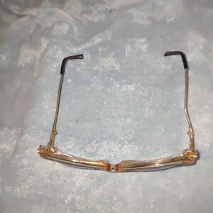 May include: A pair of spectacles featuring a translucent, light-coloured frame with a subtle golden tint. The thin metal temples are finished with black plastic tips at the ends. The glasses are positioned to show the bridge and the inner frame structure.