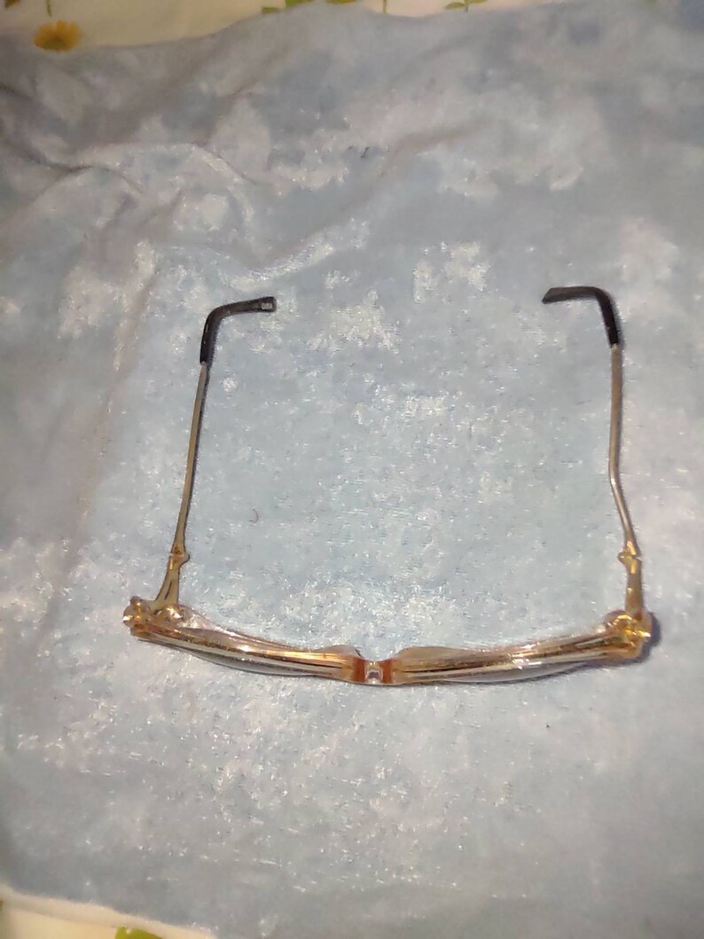 May include: A pair of spectacles featuring a translucent, light-coloured frame with a subtle golden tint. The thin metal temples are finished with black plastic tips at the ends. The glasses are positioned to show the bridge and the inner frame structure.
