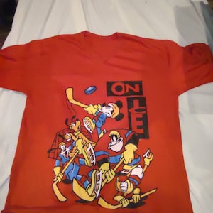 May include: Red v-neck t-shirt with a cartoon hockey scene featuring Mickey Mouse, Goofy, Donald Duck, and other characters. The phrase "ON ICE" is printed in black. The shirt is made of a soft material.