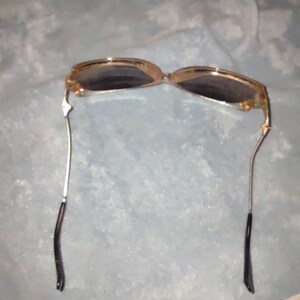 May include: Sunglasses with a translucent amber-coloured frame and dark tinted lenses. The thin metal temples are silver-toned and feature black plastic tips at the ends. The design includes a bridge connecting the two oval-shaped lens frames.