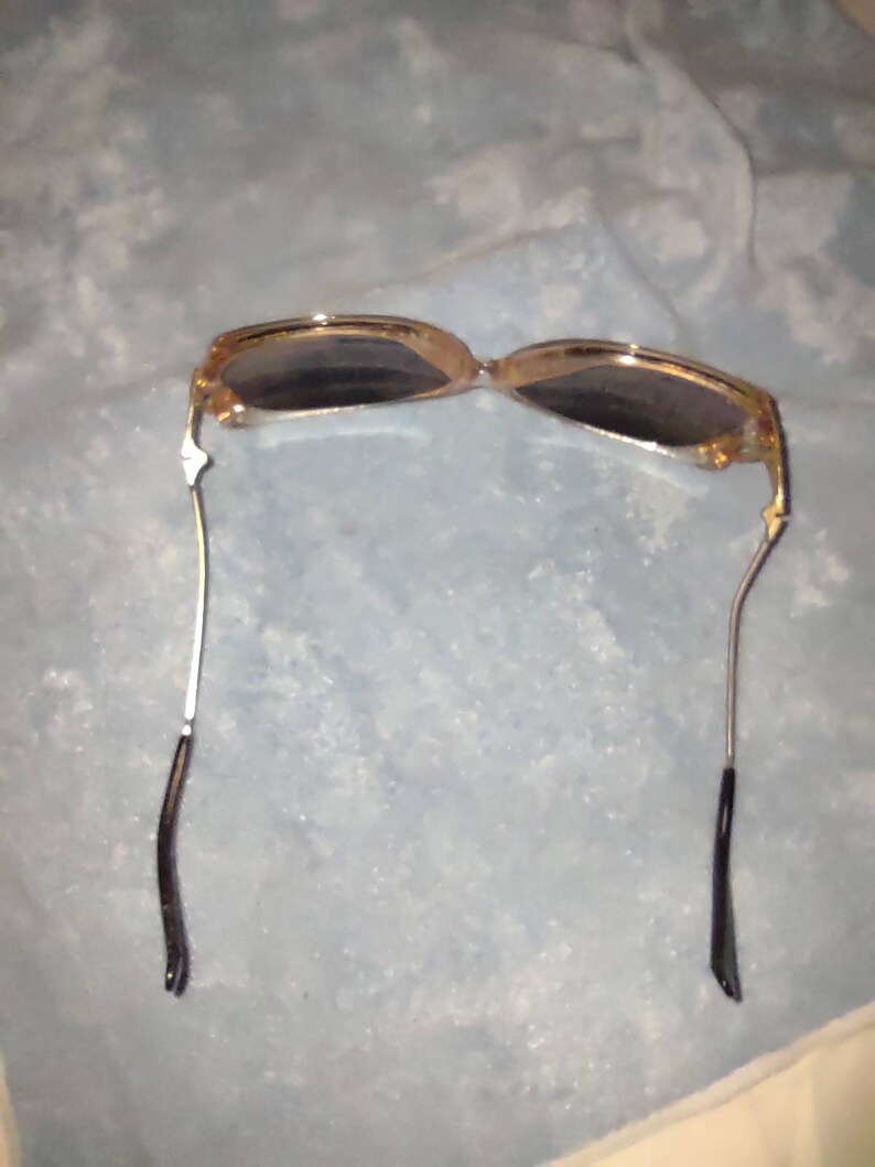 May include: Sunglasses with a translucent amber-coloured frame and dark tinted lenses. The thin metal temples are silver-toned and feature black plastic tips at the ends. The design includes a bridge connecting the two oval-shaped lens frames.