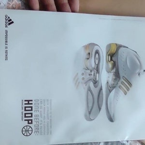 May include: A white Adidas advertisement featuring two basketball shoes. The shoes are primarily white and gray with yellow accents. The word "HOOP" is displayed in bold letters, along with the Adidas logo and the phrase "Impossible is Nothing."