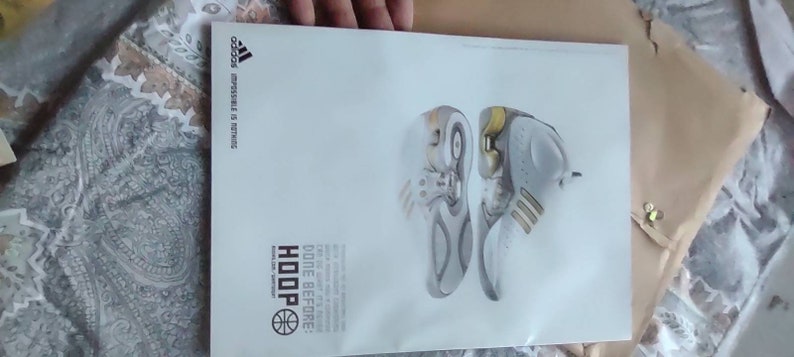 May include: A white Adidas advertisement featuring two basketball shoes. The shoes are primarily white and gray with yellow accents. The word "HOOP" is displayed in bold letters, along with the Adidas logo and the phrase "Impossible is Nothing."