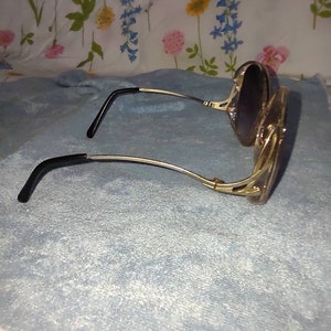 May include: Sunglasses with gold-toned metal frames and dark-tinted lenses. The temples feature a decorative cutout design and end in black plastic tips. The frames have a curved, rounded shape, and the metal components exhibit a polished finish.