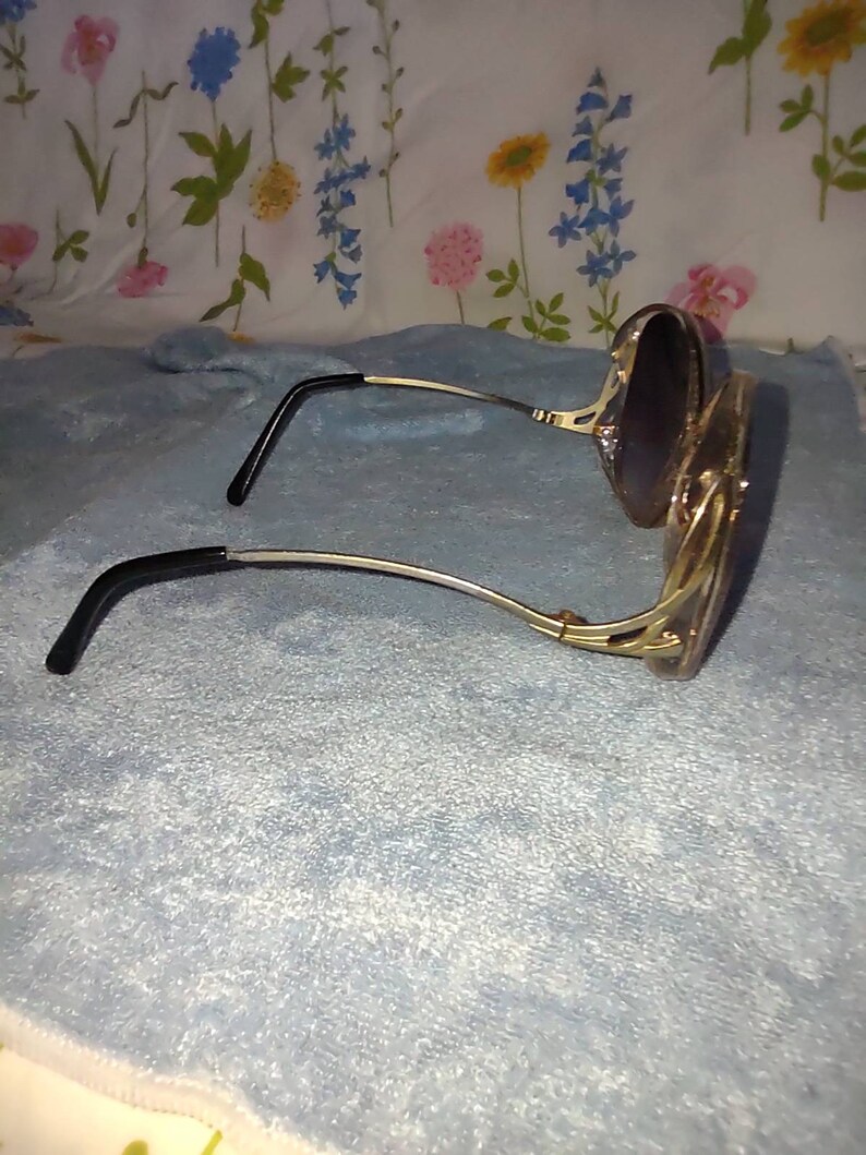 May include: Sunglasses with gold-toned metal frames and dark-tinted lenses. The temples feature a decorative cutout design and end in black plastic tips. The frames have a curved, rounded shape, and the metal components exhibit a polished finish.