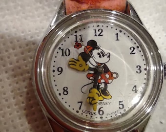 May include: A silver-toned Lorus Disney quartz watch with a white face featuring Minnie Mouse. The watch has a light pink leather strap. The watch face displays the numbers 1 through 12.