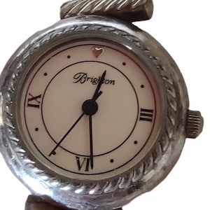 May include: Brighton silver-tone watch with a white face, black hands, and Roman numeral hour markers. The watch band features ornate, scrolling designs. The watch face reads "Brighton".