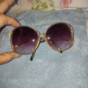 May include: Oversized sunglasses featuring a translucent, light-coloured frame with a rounded shape. The lenses have a dark purple gradient tint that transitions to a lighter shade. The thin metal temples are gold-toned and connect to the frame.