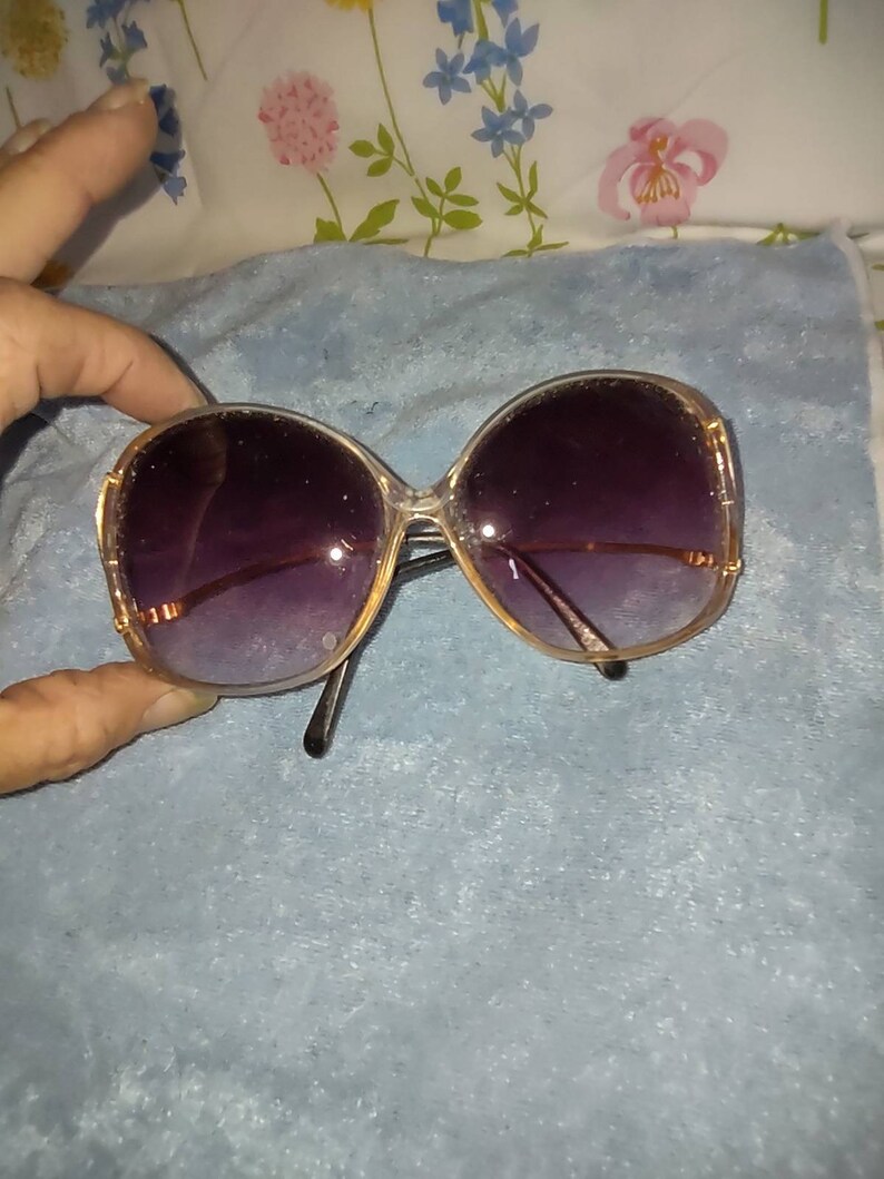 May include: Oversized sunglasses featuring a translucent, light-coloured frame with a rounded shape. The lenses have a dark purple gradient tint that transitions to a lighter shade. The thin metal temples are gold-toned and connect to the frame.