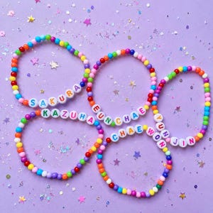 May include: Four circular bracelets made of multicolored plastic beads. Each bracelet features white letter beads spelling out the names SAKURA, KAZUHA, EUN CHAE, and YUN JIN. The beads are arranged in a repeating rainbow color pattern throughout each band.