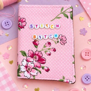 Op de afbeelding: Een rechthoekige stoffen hoes met een roze achtergrond met witte stippen en een bloemenpatroon van roze en witte bloemen met groene bladeren. De woorden STITCH BITCH zijn gespeld met kleurrijke plastic letterkralen op de voorkant.