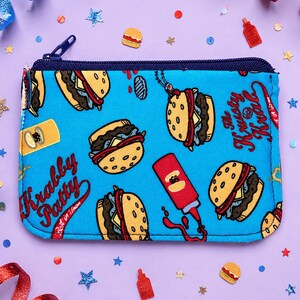 May include: A rectangular blue zippered pouch featuring a repeating pattern of Krabby Patty burgers, red ketchup bottles, and the text Krabby Patty and The Krusty Krab in red script. The pouch has a dark blue zipper closure along the top edge.