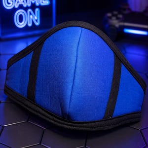 May include: A blue face mask with black trim and vertical black fabric accents. The mask features a central seam and a contoured shape, constructed from solid blue fabric with black binding along the top, bottom, and side edges for structure.