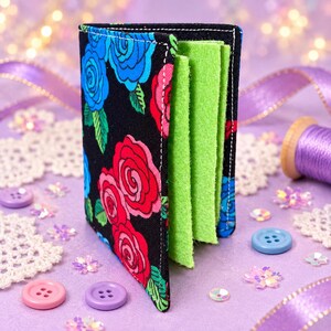 May include: A fabric needle book with a black exterior featuring a vibrant floral pattern of red and blue roses with green leaves. The interior contains multiple lime green felt pages designed for storing needles, with white stitching along the edges.