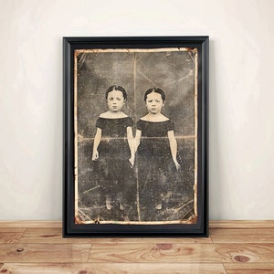 May include: Black and white vintage photograph of two young girls in dresses, standing side by side. The photo is framed in a black frame.