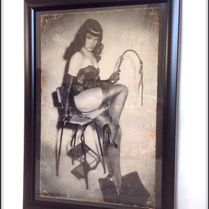 May include: A black and white photograph of a woman wearing a leather outfit, sitting on a stool and holding a whip. The photo is framed in a black frame.