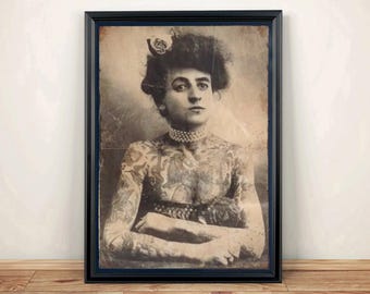 Puede incluir: Una fotografía vintage en tono sepia enmarcada de una persona con tatuajes extensos que cubren sus brazos y pecho. El individuo lleva un collar de perlas y tiene el cabello recogido con un accesorio decorativo, presentado en un marco negro.