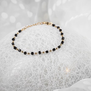 Peut inclure: Un bracelet délicat en chaîne dorée avec des perles carrées noires. Le bracelet est sur un fond blanc.
