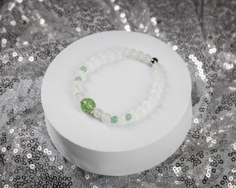 May include: A white beaded bracelet with a central green bead and smaller green accent beads. The bracelet is displayed on a white circular platform against a shimmering silver background. The bracelet features a silver clasp.