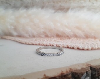May include: A silver ring with a band of small, clear gemstones. The ring is set against a soft, textured backdrop of cream and pink. The ring is displayed on a light-coloured wooden surface.