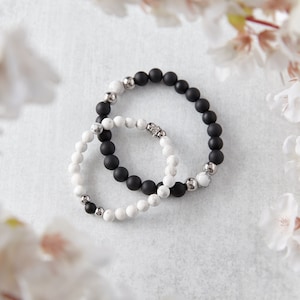 May include: Two beaded bracelets, one with black beads and the other with white beads, are displayed. The bracelets have silver-colored accents. The image is framed by white flowers, creating a soft, natural aesthetic.