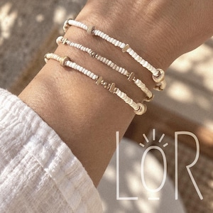 May include: Three stacked bracelets featuring small white seed beads interspersed with metallic gold-toned cylindrical and flat accent beads. The bracelets are worn together on a wrist, showcasing a delicate design with varied bead shapes.