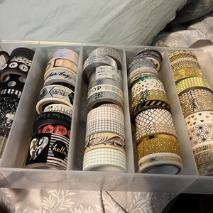 May include: A clear plastic organizer filled with decorative washi tape rolls. The tape features various patterns, including stripes, floral designs, and gold accents. Some rolls have text like "hello" and "top dream".