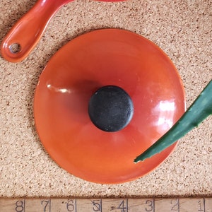 May include: Orange enameled cast iron pot lid with a black knob. The lid is sitting on a cork surface.