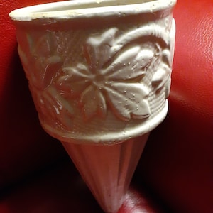 May include: Off-white ceramic wall pocket planter with a cone-shaped base. The planter features a textured floral design around the top. The planter is designed to be hung on a wall and is suitable for small plants or flowers.