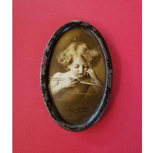 May include: An oval-shaped metal frame with a sepia-toned image of a cherub holding a bow and arrow. The image is surrounded by a dark brown border and the text "Copyright 1907 F.A. Rinehart" is visible at the bottom.