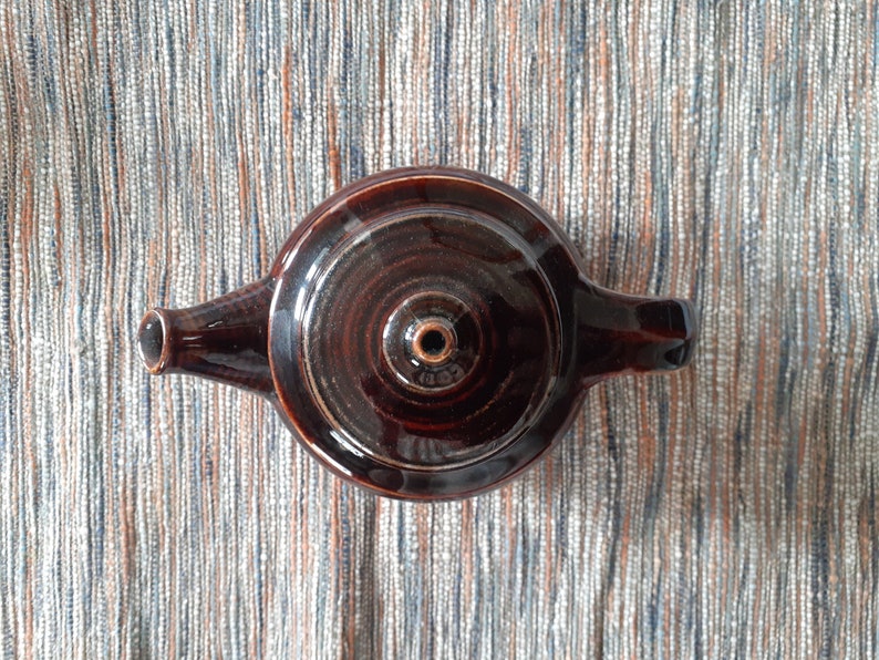 May include: A brown ceramic teapot with a round body and a single handle. The teapot has a small spout and a lid with a small hole in the center.