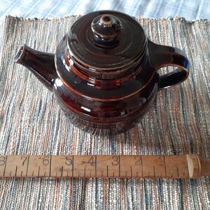 May include: A brown ceramic teapot with a lid and a handle. The teapot is sitting on a woven rug with a wooden ruler underneath.