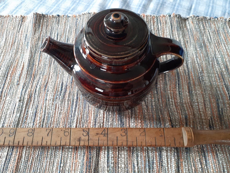 May include: A brown ceramic teapot with a lid and a handle. The teapot is sitting on a woven rug with a wooden ruler underneath.