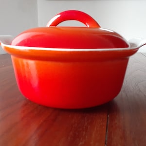 May include: Orange and red enameled cast iron pot with a white rim and lid. The pot has two small handles on the sides and a single handle on the lid.