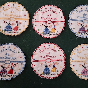 May include: Six white embroidered round coasters with scalloped edges. Each coaster features a colorful embroidered design of two women in traditional dress with the text "Recuerdo de Mallorca" in red.