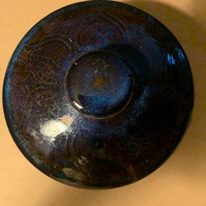 May include: A circular ceramic lid with a dark blue and black glaze featuring a subtle crackle finish. The surface displays a faint, embossed floral pattern radiating from a central knob, with reflective highlights across the glossy texture.