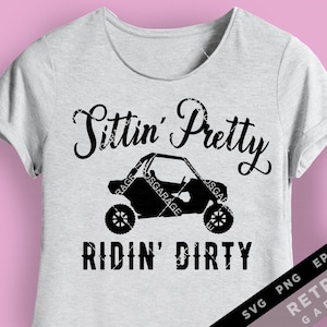 Puede incluir: Una camiseta blanca con texto negro que dice "Sittin' Pretty Ridin' Dirty". Una silueta negra de un vehículo side-by-side está en el centro de la camiseta.