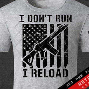 May include: A light gray t-shirt featuring a distressed American flag design with a black rifle overlay. The text "I DON'T RUN I RELOAD" is printed below the flag. An American flag is also on the sleeve.