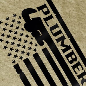 May include: A tan-coloured graphic tee featuring a distressed American flag design with a black wrench silhouette and the word "PLUMBER" in bold, angled text. The flag's stars and stripes are prominently displayed.